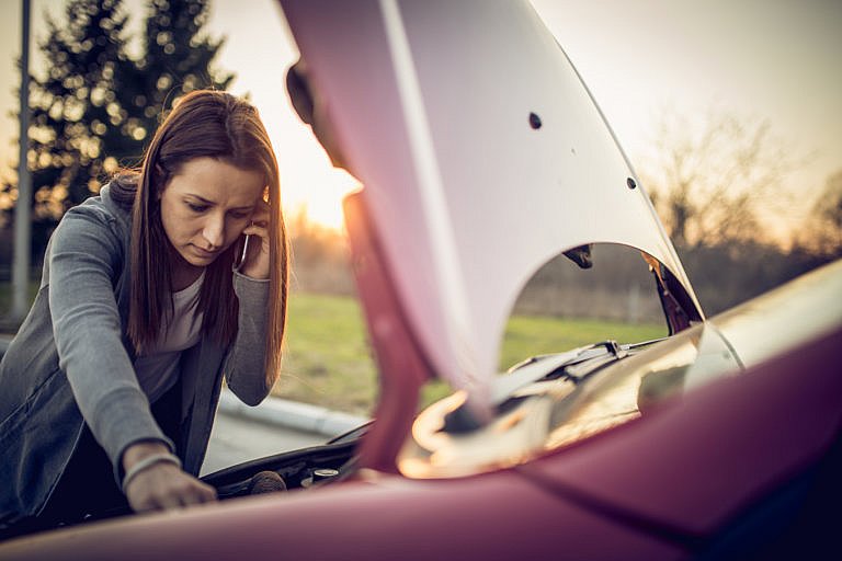 A young woman on the phone is looking under the open hood of a car with a worried expression.