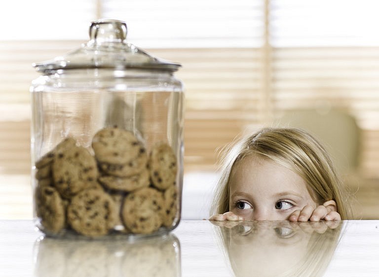 Child peeking over a counter at a jar full of cookies.