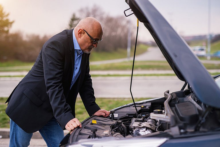 A bald man with glasses and a suit jacket is looking under the open hood of a car.