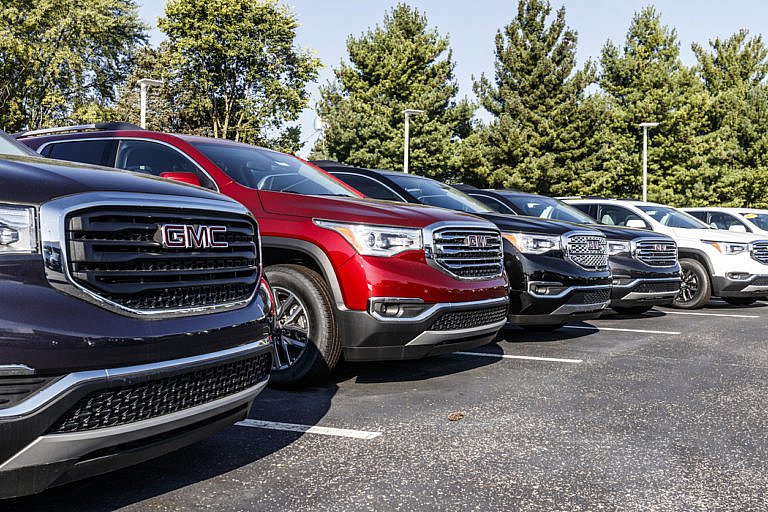 A line of new GMC SUVs is parked at a dealership, including a dark blue, a red, and a black one, with trees in the background.