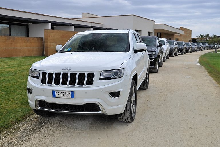 White Jeep SUV at the front of a line of cars near a modern building.