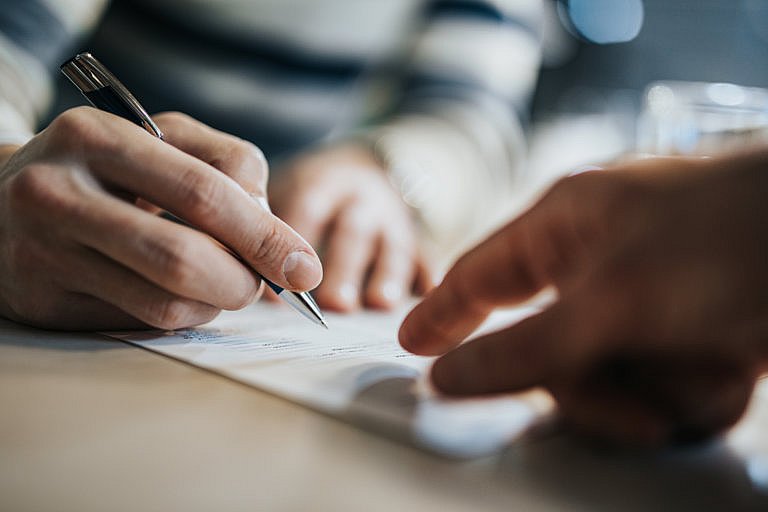 A close-up shot shows a person's hands signing a document with a pen, while another person's hand points to the paper.