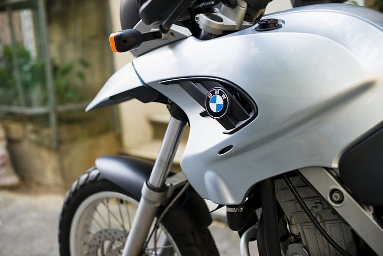 A close-up of a silver BMW motorcycle shows the logo on the front fairing and part of the front wheel and suspension.