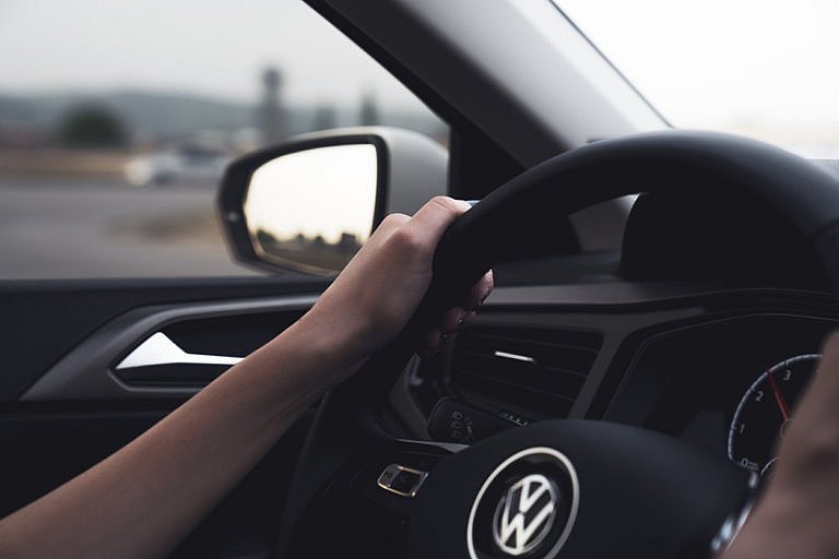 A driver's hand is on the steering wheel of a Volkswagen, with a blurred view of the road and a side mirror showing the reflection of the landscape.