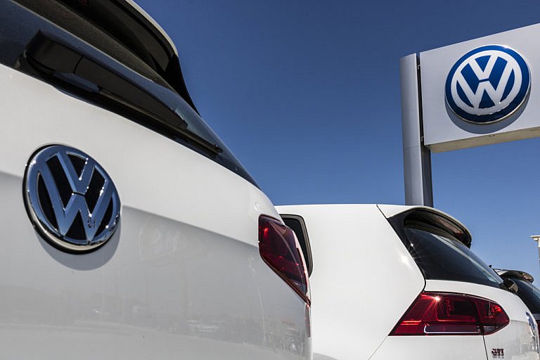 A view from behind two white Volkswagen Golf GTI cars shows the VW logo and a large sign with the VW logo on a sunny day.