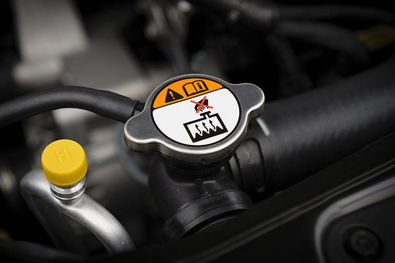 A close-up of a car's engine shows a radiator cap with a warning label on it, advising not to open it while hot.