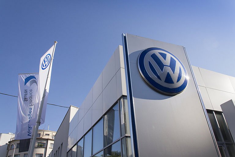 A low-angle view of a modern Volkswagen dealership building with a large logo on the corner and a flag flying in the foreground.