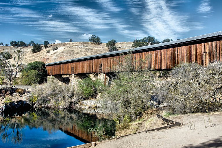 Wooden covered bridge crossing a calm river with hills in the background.