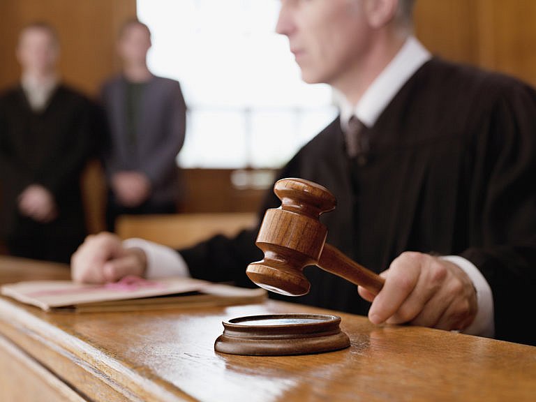 A judge in a black robe is holding a gavel over a sound block at a wooden bench in a courtroom. Two other people stand blurred in the background.