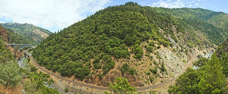 Railroad track winding through a forested mountain canyon with a bridge in the distance.