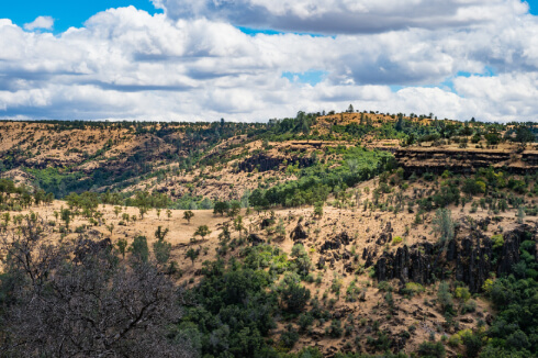 Scenic canyon landscape with rolling hills, trees, and partly cloudy sky.