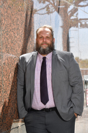 A bearded man in a grey suit and dark tie stands with his hands in his pockets against a red-brown marble wall.
