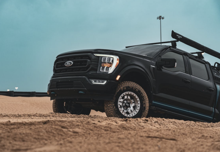 A black Ford F-150 drives through sand with muddy tires.