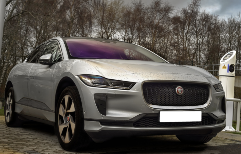 A silver Jaguar I-Pace electric SUV is parked and charging on a cloudy day.