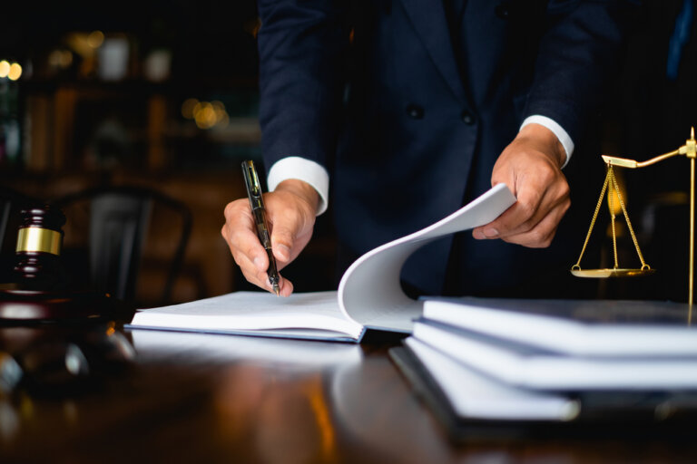 A person in a suit is signing a document on a desk with a gavel and a scale of justice nearby.