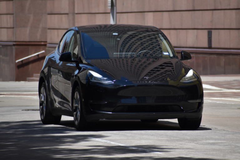 A shiny black Tesla Model Y is on a city street, with the reflection of a tall building visible on its windshield.