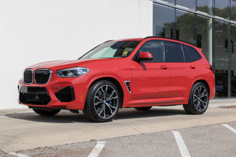 A **shiny red BMW X3 M SUV** is parked outside a modern building with large glass windows.