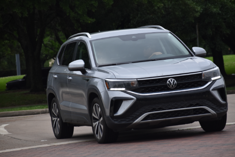 A silver Volkswagen Taos is driving on a paved road, turning toward the camera with trees in the background.