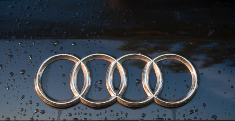 A close-up of a wet black car shows the chrome Audi logo, which consists of four interlocking rings.
