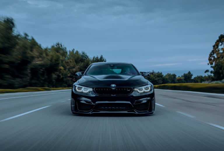 A low-angle, front-facing shot of a sleek black BMW M4 driving down a road with trees and a cloudy sky in the background.