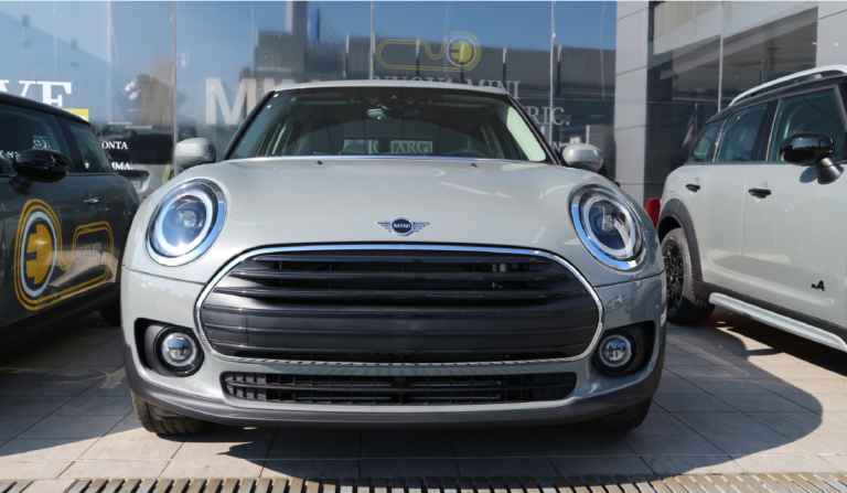 A silver Mini Cooper is parked in the middle of two other vehicles, with a dealership building behind it.