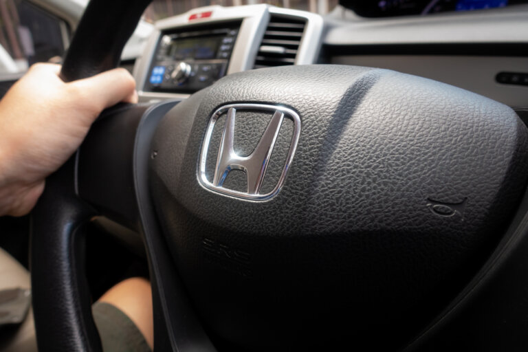 A driver's hand rests on the steering wheel of a Honda vehicle, featuring the brand's chrome emblem.
