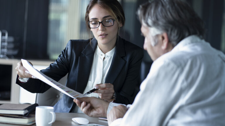A woman in business attire is showing and explaining a document to an older man in a meeting setting.
