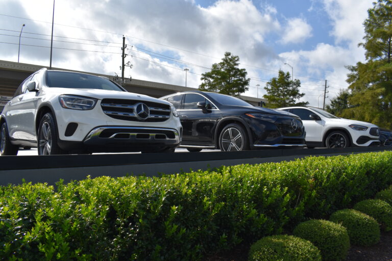 A row of Mercedes-Benz SUVs is on display, with a white one in the foreground and a black one and a white one behind it. The cars are positioned above a line of hedges.