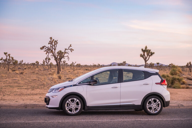 A white Chevrolet Bolt electric vehicle is parked on the side of a road in a desert landscape with Joshua trees.