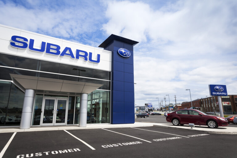 A **Subaru** dealership building has a large logo on the side and a red sedan parked in front.