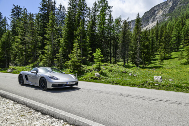 A silver Porsche Boxster convertible is driving on a scenic road with a forest and mountains in the background.