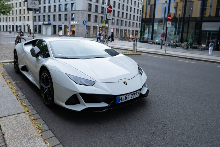 A low-angle view of a white Lamborghini Huracán is parked on a city street with pedestrians and buildings in the background.