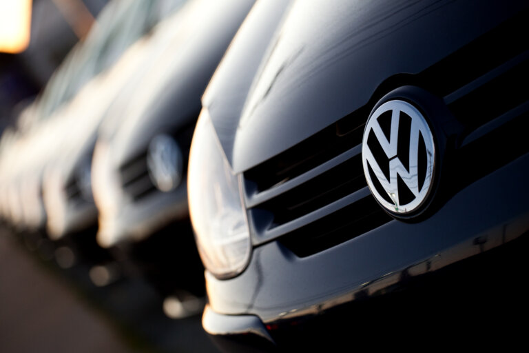 A line of new, dark-colored Volkswagen cars is shown, with a close-up on the logo and grille of the car in the foreground.
