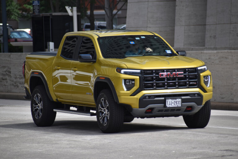 A gold GMC Canyon AT4X pickup truck is parked on a paved lot in front of a modern building.