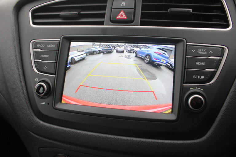 The infotainment screen of a car shows a backup camera view of several cars parked in a lot. Red and yellow lines are overlaid on the image to assist with parking.