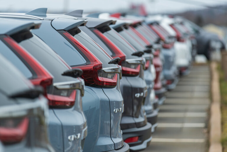 A **long line of new, gray SUVs** is parked outside a car dealership. The image focuses on the rear lights of the vehicles, with a shallow depth of field.