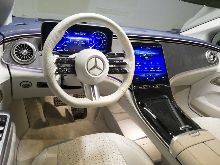 An interior view of a modern Mercedes-Benz shows the steering wheel and the sleek, wide digital dashboard.