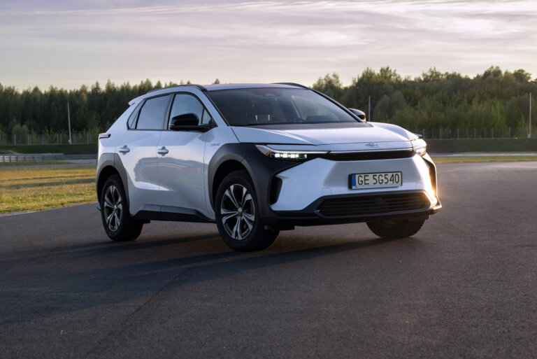 A white Toyota bZ4X electric SUV is parked on an asphalt road with a forest in the background.