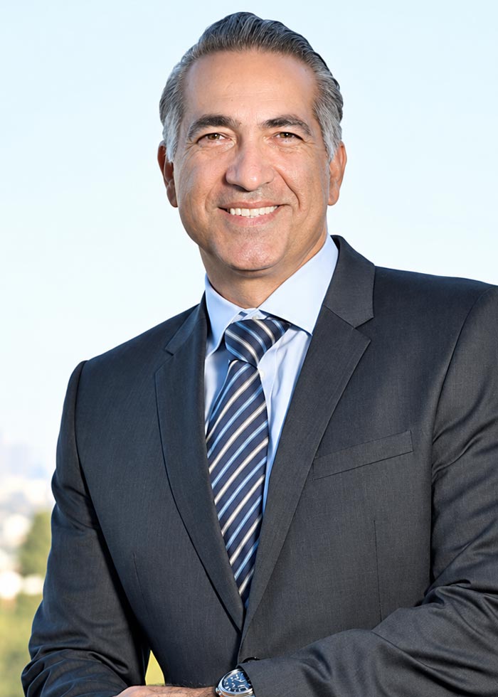 Sepehr in a dark suit, light blue shirt, and striped tie, posed against a pale background.