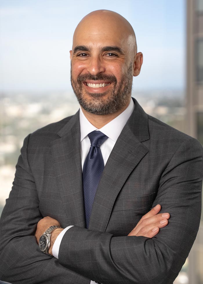 Smiling bald man with a beard wearing a dark suit and tie, posed with folded arms against a blurred cityscape.