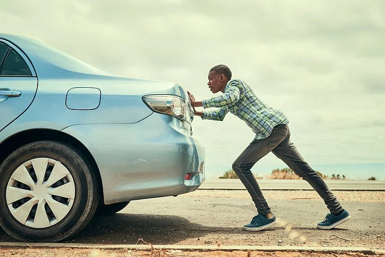 A man is pushing a blue car on the side of the road.