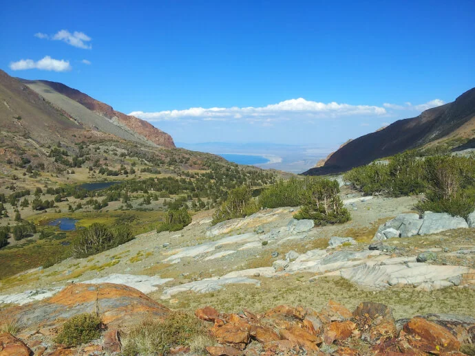 A scenic mountain valley with rocky terrain, green trees, and a distant lake under blue skies.