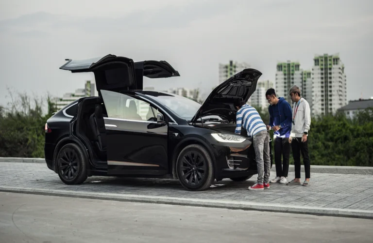 Black Tesla Model X with open falcon doors and hood being inspected by three people.
