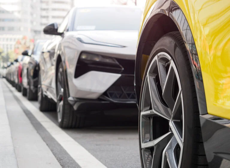 A row of cars is parked on a city street, with the front of a yellow car and its wheel in the foreground.