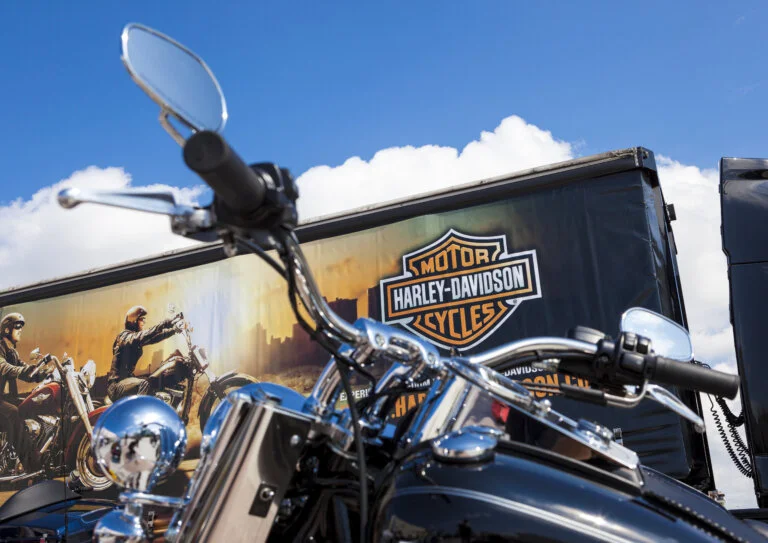 A **Harley-Davidson** motorcycle is in the foreground, with its handlebars and chrome details in focus. Behind it is a semi-truck trailer with the Harley-Davidson logo and graphics.