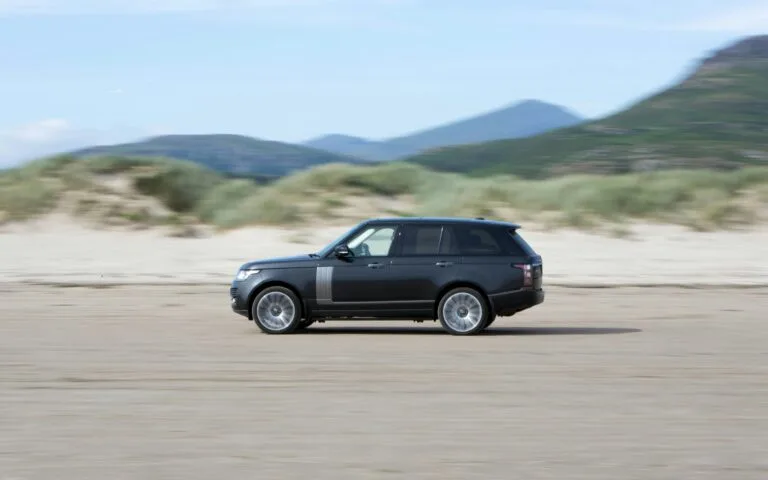 Dark Land Rover SUV driving across sandy terrain, with dunes and mountains in the background.