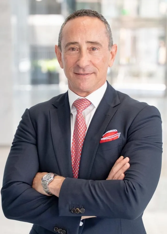 Portrait of a man in a dark suit and red tie, standing with his arms crossed.